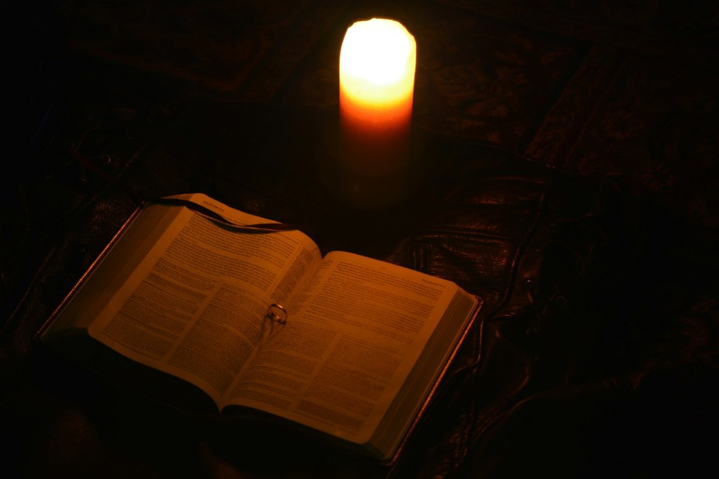 A lit candle burning beside an open old book in low light, evoking the historical records of the Salem witch trials