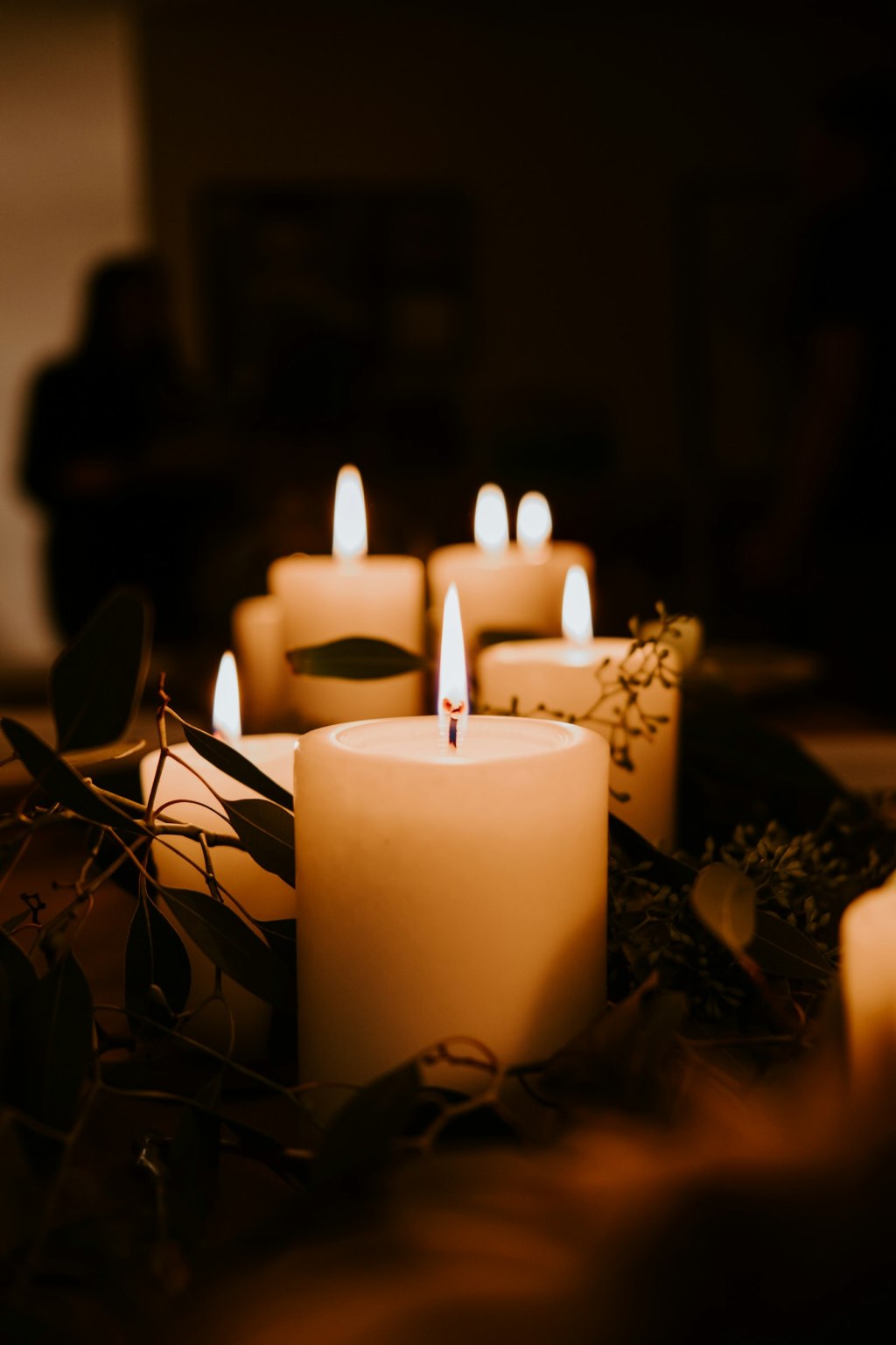 Lit white pillar candles surrounded by green eucalyptus leaves in a dark room, a simple witch altar setup